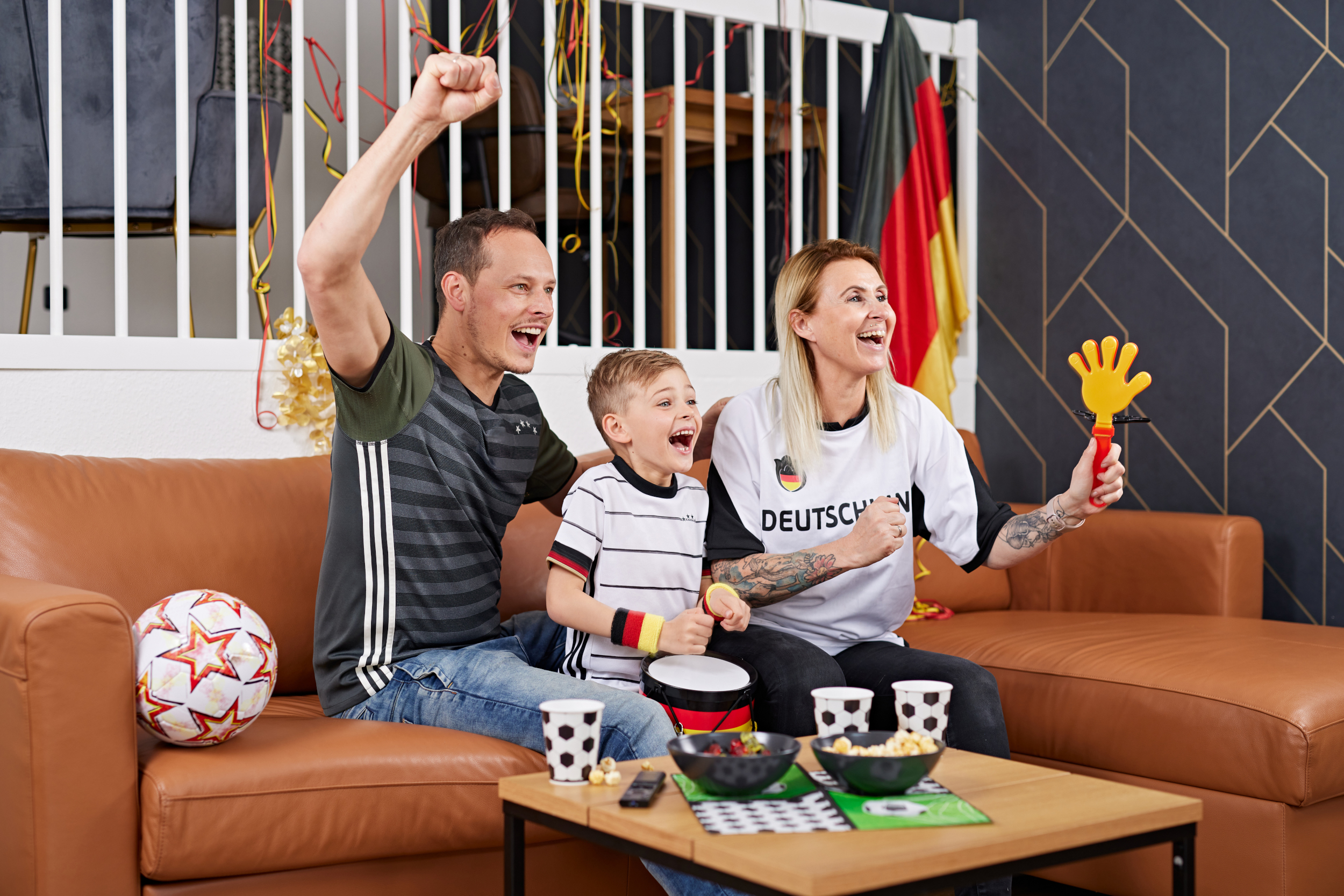 Papa, Mama und Kind sitzen auf dem Sofa und jubeln Deutschland zu